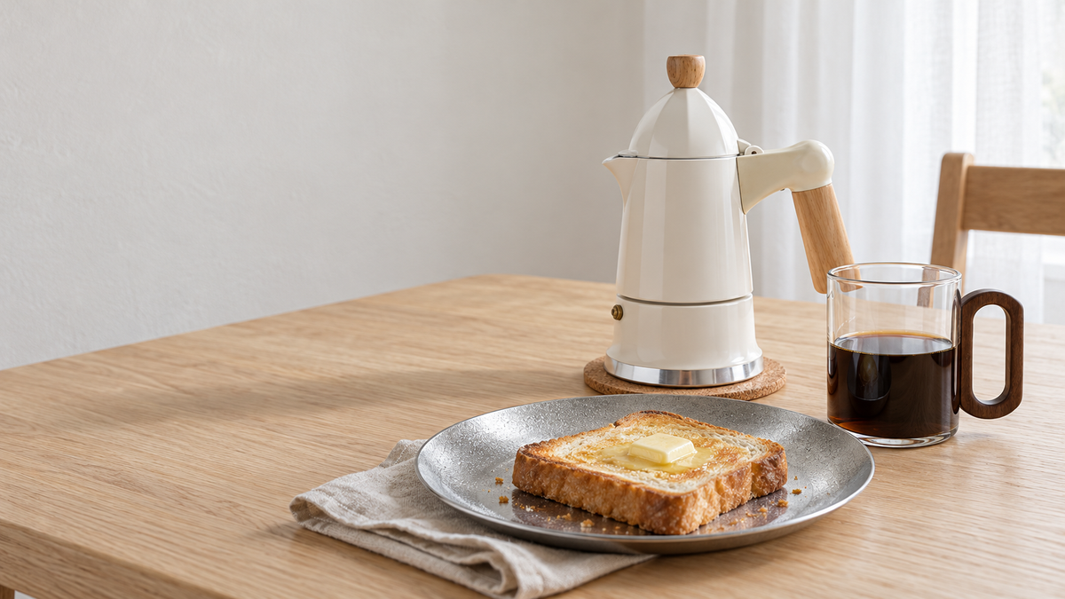 柔らかな自然光の朝の食卓。白い木製ハンドルのモカポット、コーヒー入りの持ち手付きガラスマグ、トーストを載せたシルバーのステンレスプレート。